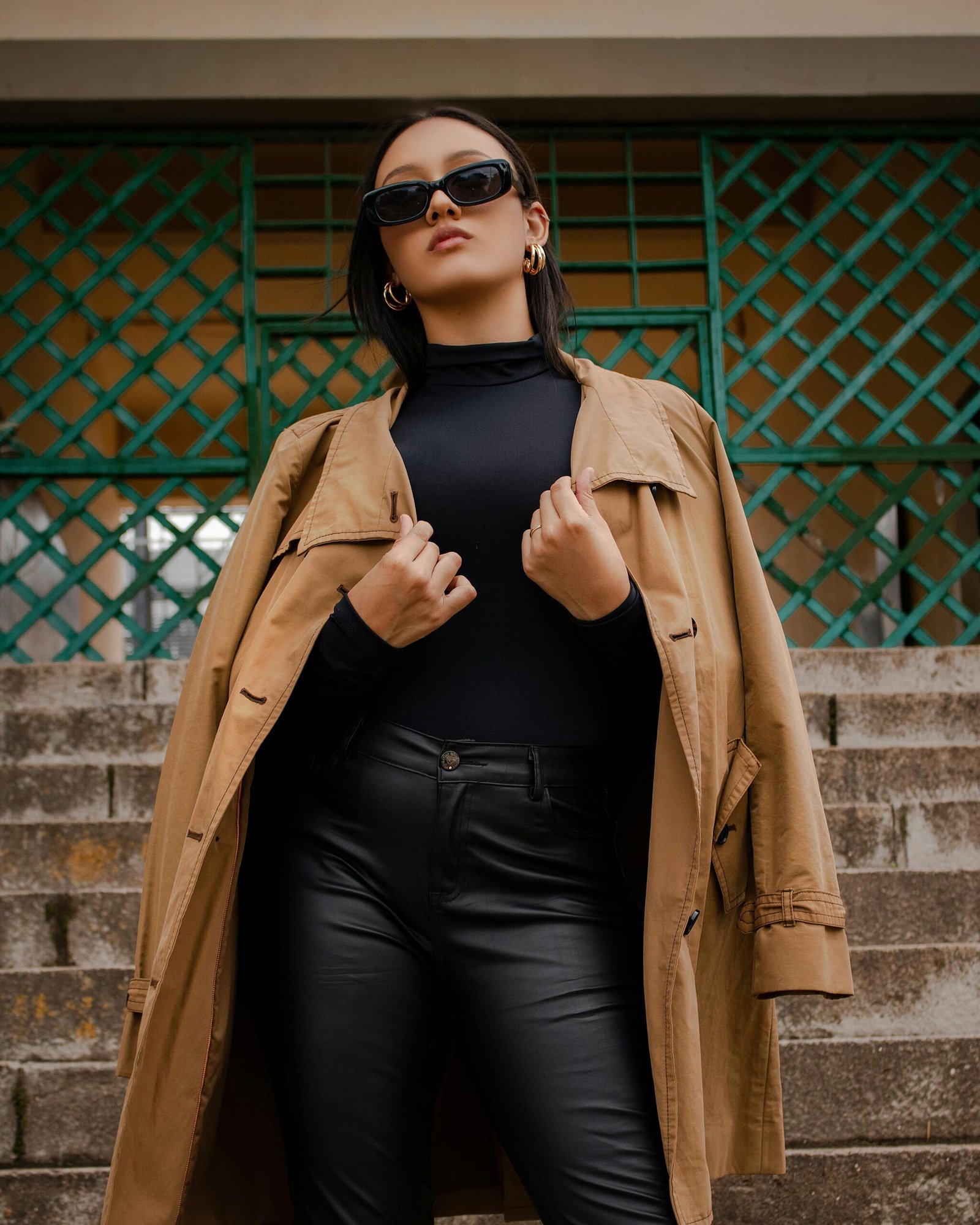 Home Fashionable woman with sunglasses poses confidently on city stairs, wearing a trendy trench coat.